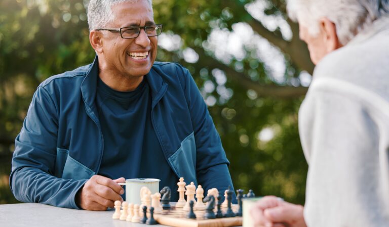 Two older men playing a game of chess at a park table, one smiling while holding a mug on the table