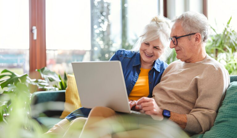 Smiling senior couple sitting on a couch and using a laptop together in a sunlit living room.