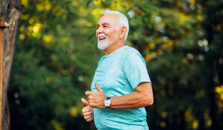 Smiling elderly man in a light blue shirt runs outdoors in a sunlit park.