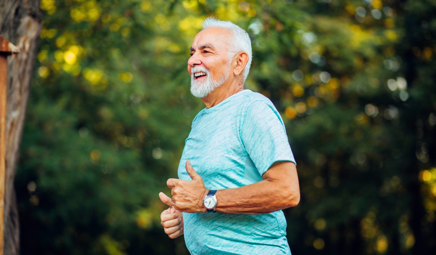 Smiling elderly man in a light blue shirt runs outdoors in a sunlit park.