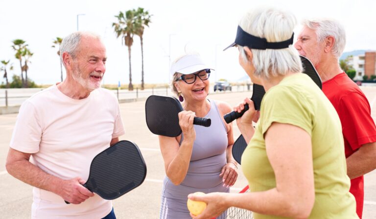 Group of seniors chatting and laughing while holding paddle tennis paddles on a sunny boardwalk.